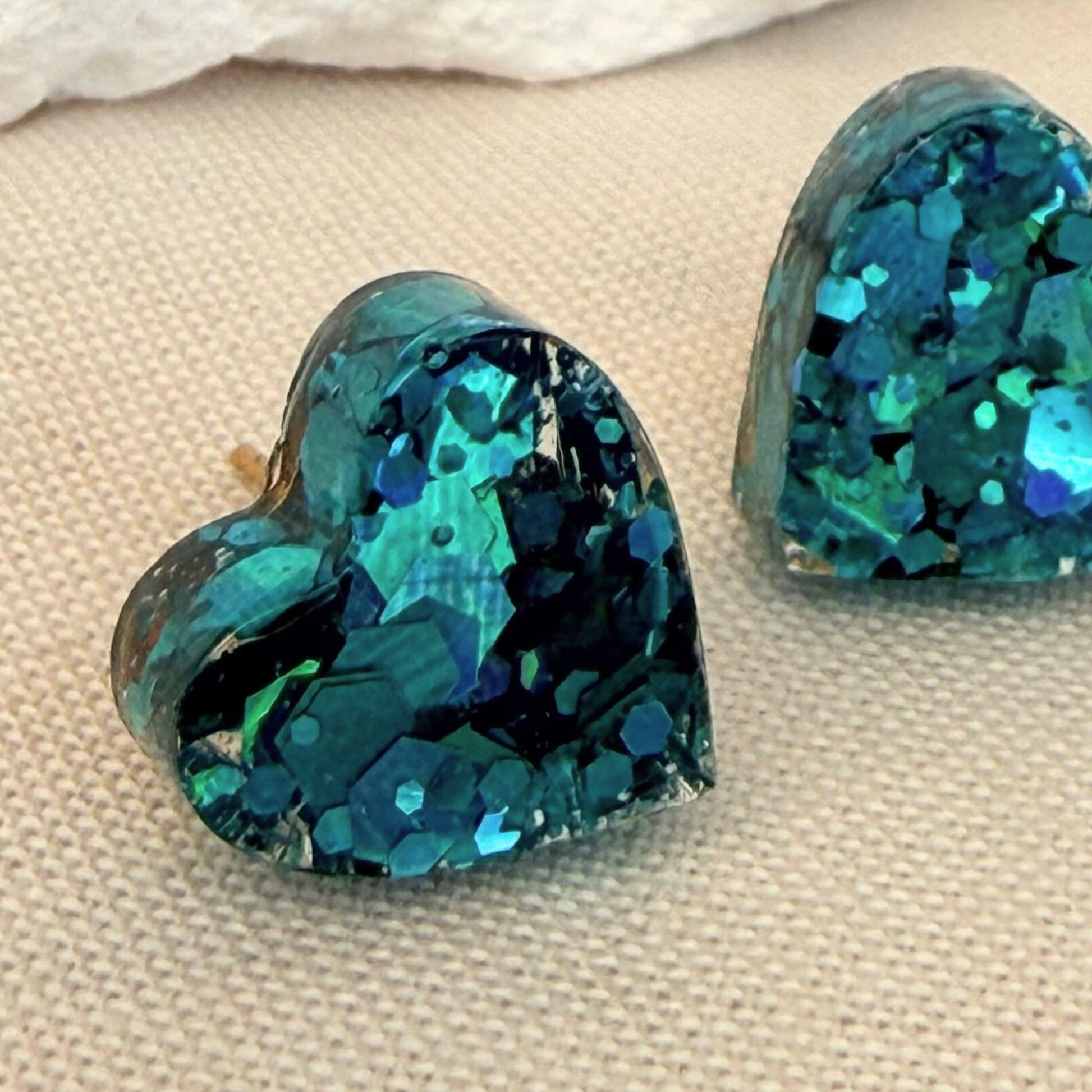 Heart-shaped resin stud earrings with teal glitter finish on a beige fabric background.