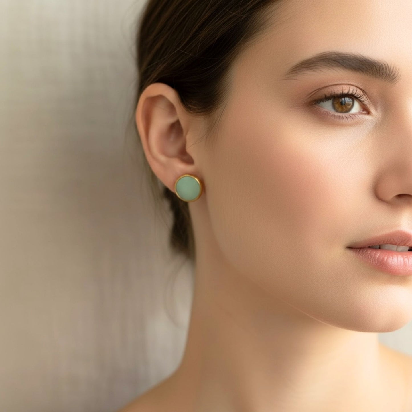 Close-up of a woman wearing Australian handmade light eucalypt green earrings with gold bezel against a neutral background.