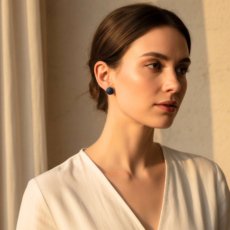 Woman wearing small round navy blue stud earrings in warm natural light in a neutral indoor setting.