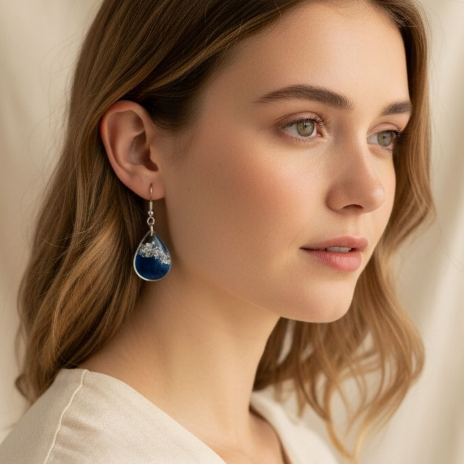 Close-up of a woman wearing a blue resin teardrop earring with silver toned hardware and details against a neutral background.
