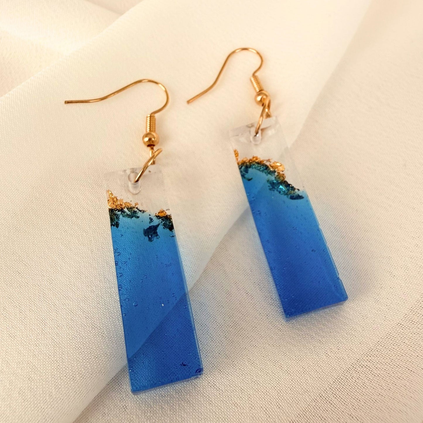 Tidal Bar handmade blue resin dangle earrings with gold leaf details and gold findings resting on white fabric.