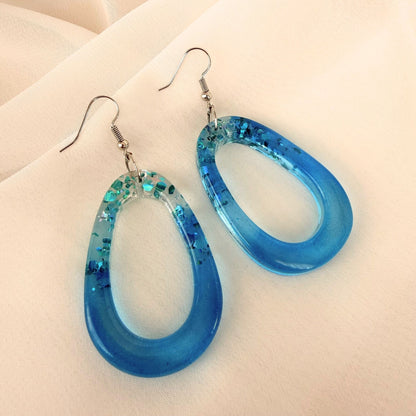 Tidal Glow oval shaped resin earrings in blue with subtle shimmer resting on white fabric.