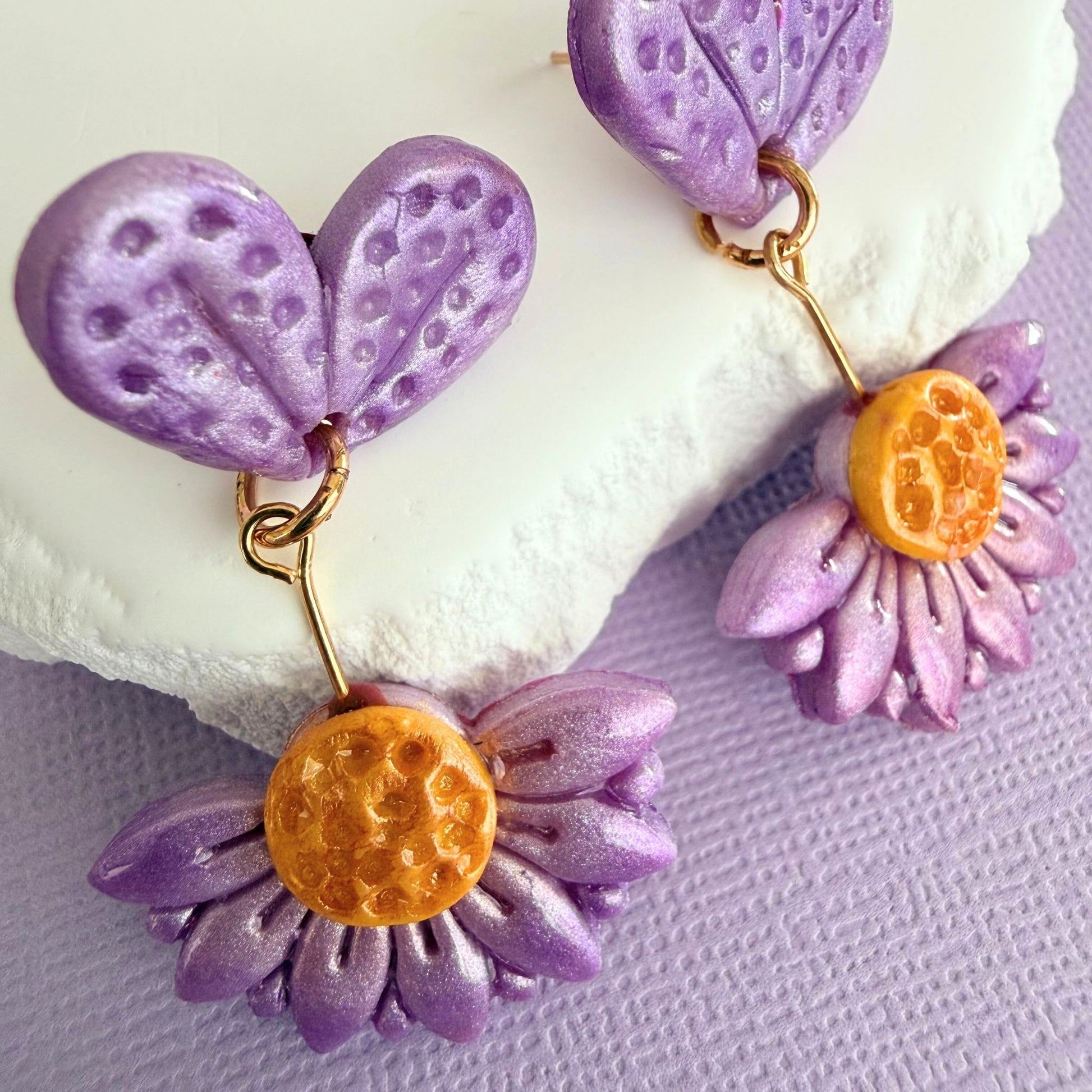 Close up of Violet Bloom floral drop earrings in purple with gold accents resting against a white stone surface on a purple background.