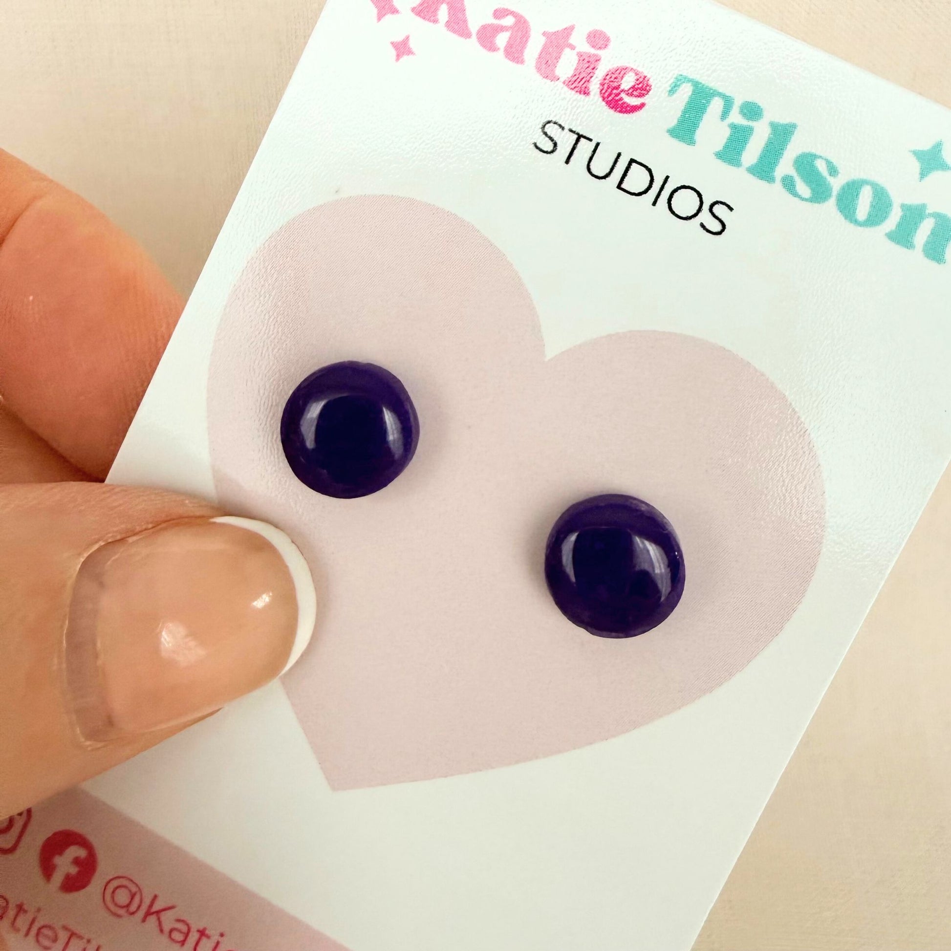 Deep violet purple circular studs on a branded earring card held by a hand.